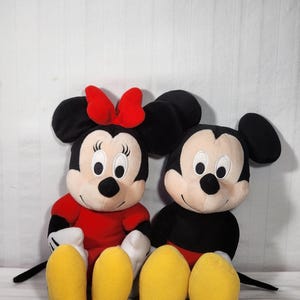 Pu&ograve; includere: Due bambole di peluche dei personaggi Disney, Topolino e Minnie, sedute fianco a fianco. Minnie indossa un vestito e un fiocco rossi, mentre Topolino ha pantaloncini neri. Entrambi hanno grandi orecchie nere, facce bianche e piedi gialli.