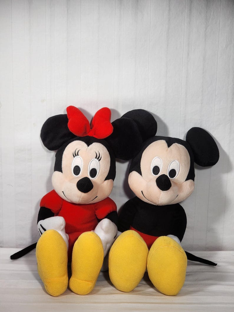 Pu&ograve; includere: Due bambole di peluche dei personaggi Disney, Topolino e Minnie, sedute fianco a fianco. Minnie indossa un vestito e un fiocco rossi, mentre Topolino ha pantaloncini neri. Entrambi hanno grandi orecchie nere, facce bianche e piedi gialli.