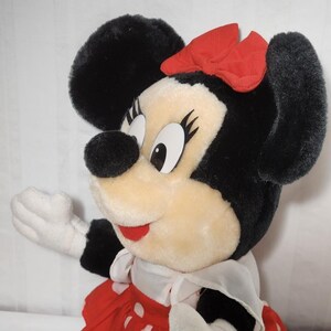 May include: A plush Minnie Mouse doll with black fur, large black ears, and a red bow. She wears a red dress with white polka dots, a white collar, and white gloves. The doll has a smiling expression and is a classic Disney character.