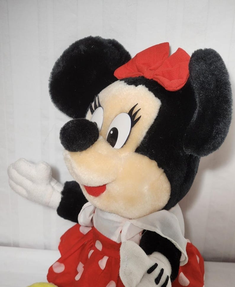 May include: A plush Minnie Mouse doll with black fur, large black ears, and a red bow. She wears a red dress with white polka dots, a white collar, and white gloves. The doll has a smiling expression and is a classic Disney character.