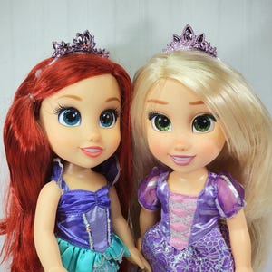 May include: Two dolls, one with red hair and the other with blonde hair, both wearing purple tiaras and dresses. The red-haired doll has a blue and purple outfit, while the blonde doll wears a purple dress with pink accents. Both dolls have large eyes.