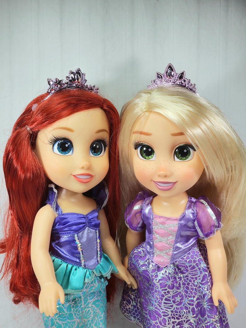 May include: Two dolls, one with red hair and the other with blonde hair, both wearing purple tiaras and dresses. The red-haired doll has a blue and purple outfit, while the blonde doll wears a purple dress with pink accents. Both dolls have large eyes.