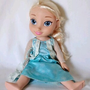 Op de afbeelding: Een blonde pop die een blauwe jurk draagt met een wit sneeuwvlokkenpatroon. De pop heeft blauwe ogen en een lichtroze huidskleur. De pop zit op een wit oppervlak.
