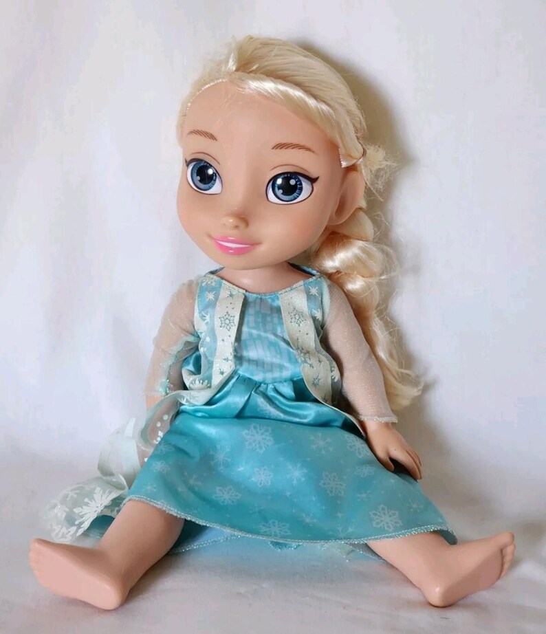 Op de afbeelding: Een blonde pop die een blauwe jurk draagt met een wit sneeuwvlokkenpatroon. De pop heeft blauwe ogen en een lichtroze huidskleur. De pop zit op een wit oppervlak.