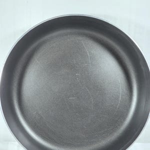 Może przedstawiać: Ciemnoszara patelnia z powłoką nieprzywierającą i srebrną obwódką. Okrągła patelnia ma lekko porysowane wnętrze. Jest pusta i gotowa do użycia.