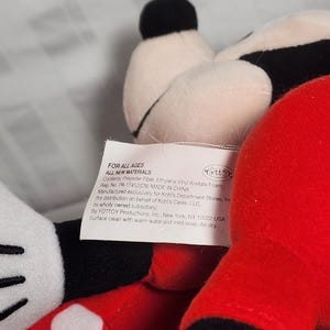 Pu&ograve; includere: Un peluche con una testa nera e bianca, un orecchio nero e un corpo rosso. Il giocattolo ha un guanto bianco con un punto bianco. Un'etichetta &egrave; visibile con il testo "FOR ALL AGES" e altri dettagli.