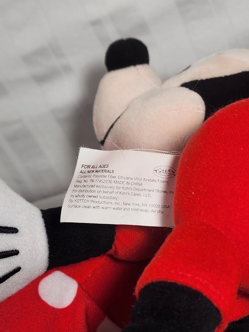 Pu&ograve; includere: Un peluche con una testa nera e bianca, un orecchio nero e un corpo rosso. Il giocattolo ha un guanto bianco con un punto bianco. Un'etichetta &egrave; visibile con il testo "FOR ALL AGES" e altri dettagli.