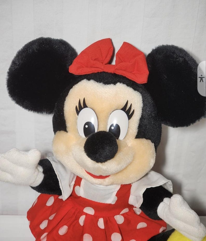 May include: A plush Minnie Mouse doll with black ears, a red bow, and a red dress with white polka dots. The doll has white gloves and a smiling expression. A classic Disney character.