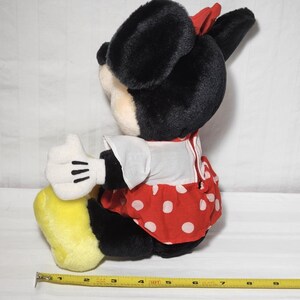 May include: A plush Minnie Mouse doll, featuring black fur, a red polka-dot dress, and a red bow. The doll has white gloves and yellow shoes. A measuring tape is visible, showing inches.