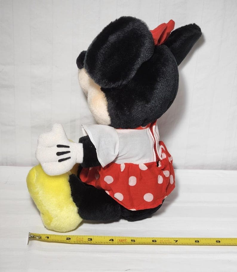 May include: A plush Minnie Mouse doll, featuring black fur, a red polka-dot dress, and a red bow. The doll has white gloves and yellow shoes. A measuring tape is visible, showing inches.
