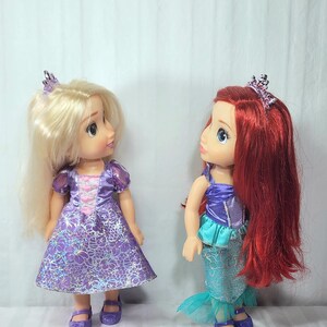 May include: Two dolls, one with blonde hair and a purple dress, the other with red hair and a mermaid tail. Both dolls wear tiaras and purple shoes. The blonde doll's dress has a floral pattern.