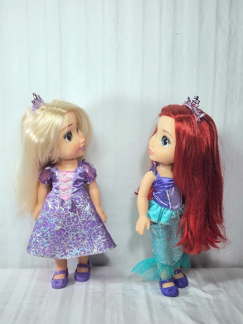 May include: Two dolls, one with blonde hair and a purple dress, the other with red hair and a mermaid tail. Both dolls wear tiaras and purple shoes. The blonde doll's dress has a floral pattern.