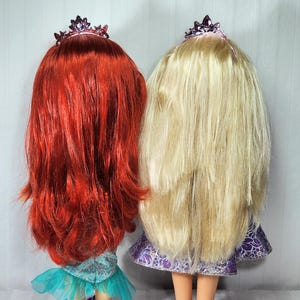 May include: Two dolls with long hair and tiaras stand side-by-side. One doll has bright red hair, a teal dress, and purple shoes. The other doll has long blonde hair, a purple dress, and purple shoes. Both dolls are facing away from the camera.