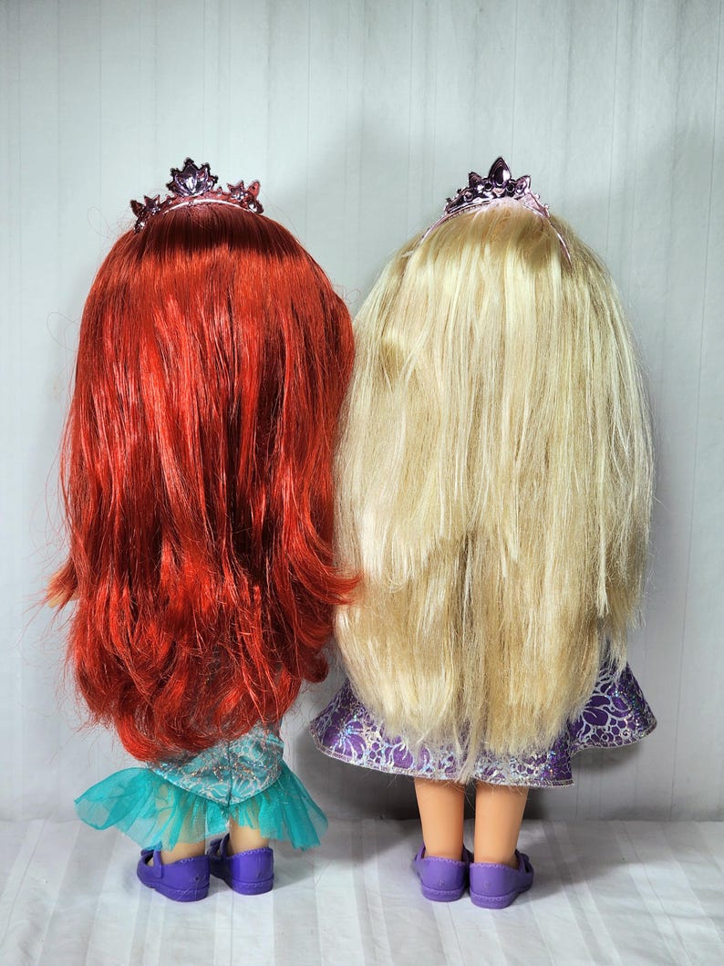 May include: Two dolls with long hair and tiaras stand side-by-side. One doll has bright red hair, a teal dress, and purple shoes. The other doll has long blonde hair, a purple dress, and purple shoes. Both dolls are facing away from the camera.