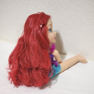 Puede incluir: Mu&ntilde;eca con largo cabello rojo, ojos azules y tez clara. La mu&ntilde;eca lleva un top morado y pantalones cortos azules. La mu&ntilde;eca est&aacute; sentada con una pierna extendida.