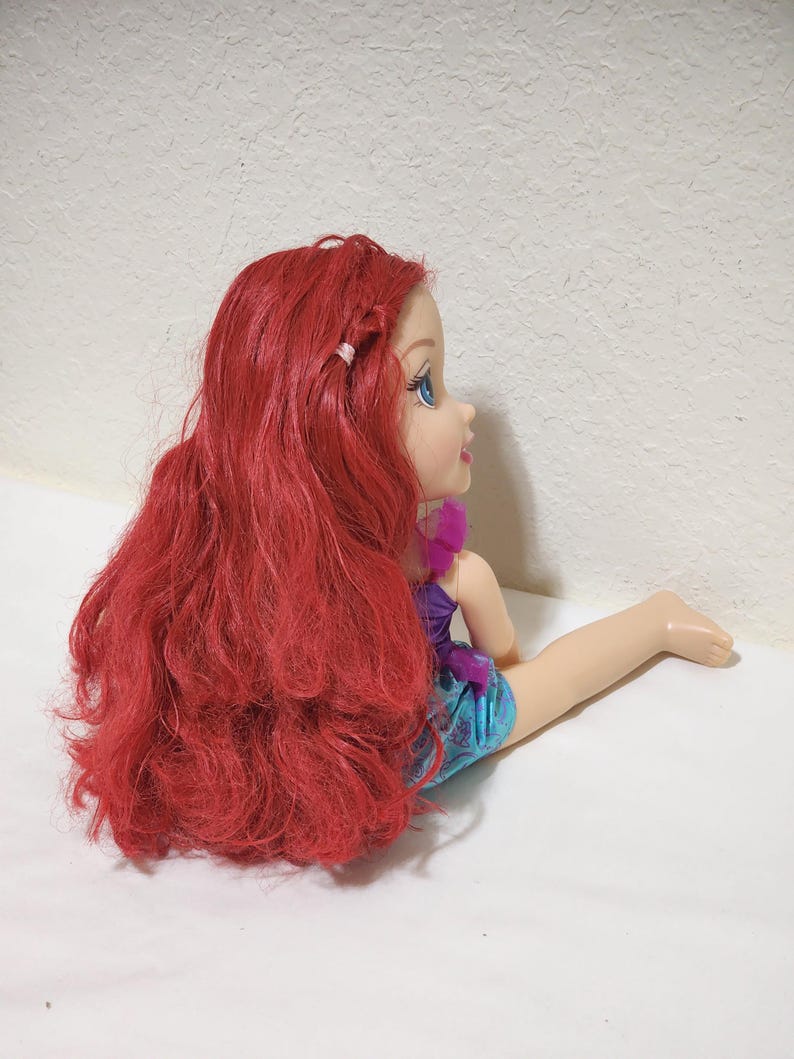 Puede incluir: Mu&ntilde;eca con largo cabello rojo, ojos azules y tez clara. La mu&ntilde;eca lleva un top morado y pantalones cortos azules. La mu&ntilde;eca est&aacute; sentada con una pierna extendida.