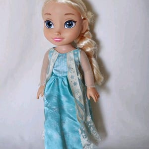 Op de afbeelding: Een pop met een blauwe en witte jurk met een sneeuwvlokkenpatroon. De pop heeft blond haar in een vlecht en blauwe ogen.