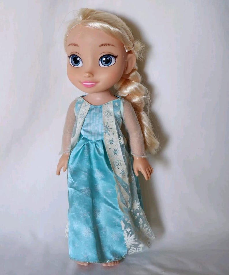 Op de afbeelding: Een pop met een blauwe en witte jurk met een sneeuwvlokkenpatroon. De pop heeft blond haar in een vlecht en blauwe ogen.