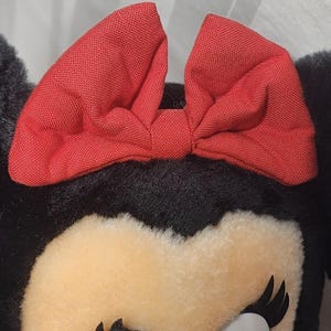 May include: Close-up of a plush toy featuring a red bow and black ears. The toy has a cream-colored face with black eyelashes and a white eye. The bow is made of a textured fabric.