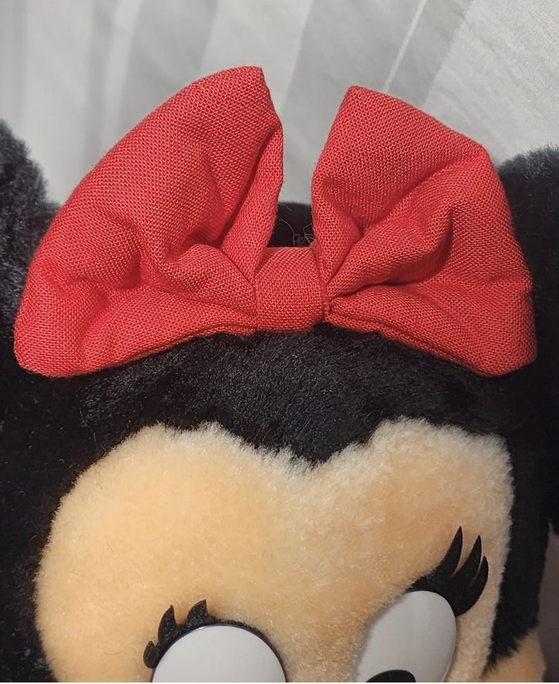 May include: Close-up of a plush toy featuring a red bow and black ears. The toy has a cream-colored face with black eyelashes and a white eye. The bow is made of a textured fabric.