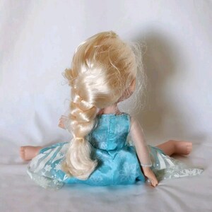 Op de afbeelding: Een blonde pop die een blauwe jurk draagt met een sneeuwvlokkenpatroon. De pop heeft een lange vlecht en zit op een wit oppervlak.