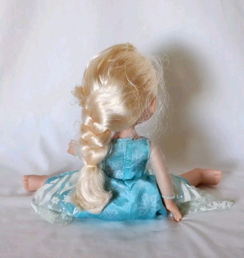 Op de afbeelding: Een blonde pop die een blauwe jurk draagt met een sneeuwvlokkenpatroon. De pop heeft een lange vlecht en zit op een wit oppervlak.