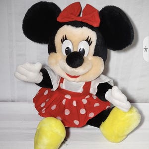 May include: A plush Minnie Mouse doll with black ears, a red bow, and a red dress with white polka dots. The doll has white gloves and yellow shoes. The doll is a popular character from Disney.