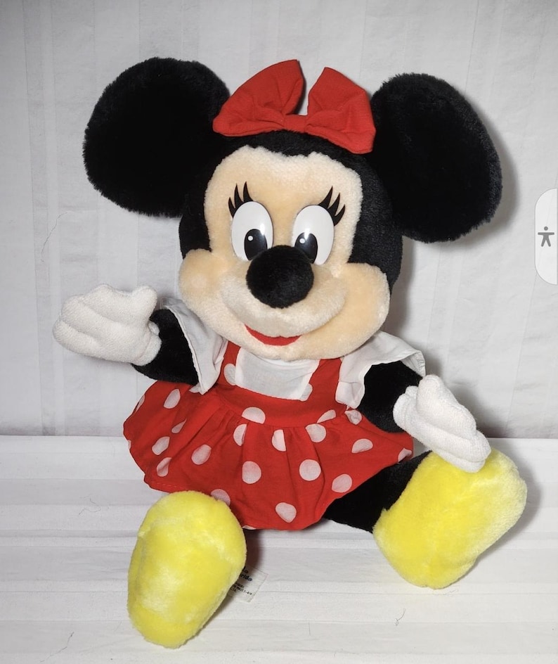 May include: A plush Minnie Mouse doll with black ears, a red bow, and a red dress with white polka dots. The doll has white gloves and yellow shoes. The doll is a popular character from Disney.