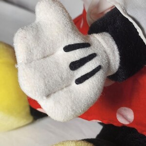 May include: Close-up of a white plush glove with three black lines on the palm. The glove is part of a plush toy, with a red polka-dot dress and black sleeve visible. A yellow object is in the lower left corner.
