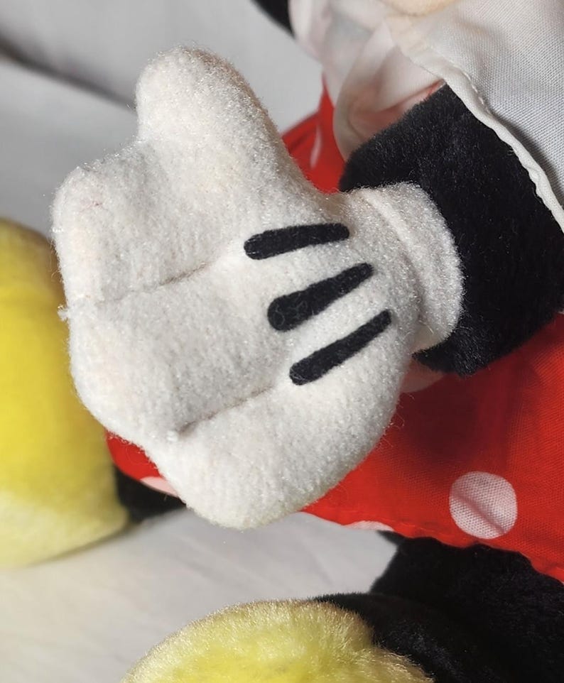 May include: Close-up of a white plush glove with three black lines on the palm. The glove is part of a plush toy, with a red polka-dot dress and black sleeve visible. A yellow object is in the lower left corner.