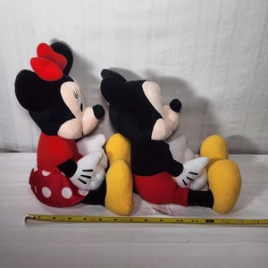 Pu&ograve; includere: Due bambole di peluche dei personaggi Disney, Minnie Mouse e Topolino, sono sedute fianco a fianco. Minnie indossa un vestito rosso a pois bianchi e un fiocco rosso. Topolino indossa pantaloncini rossi. Entrambi hanno orecchie nere e guanti bianchi.