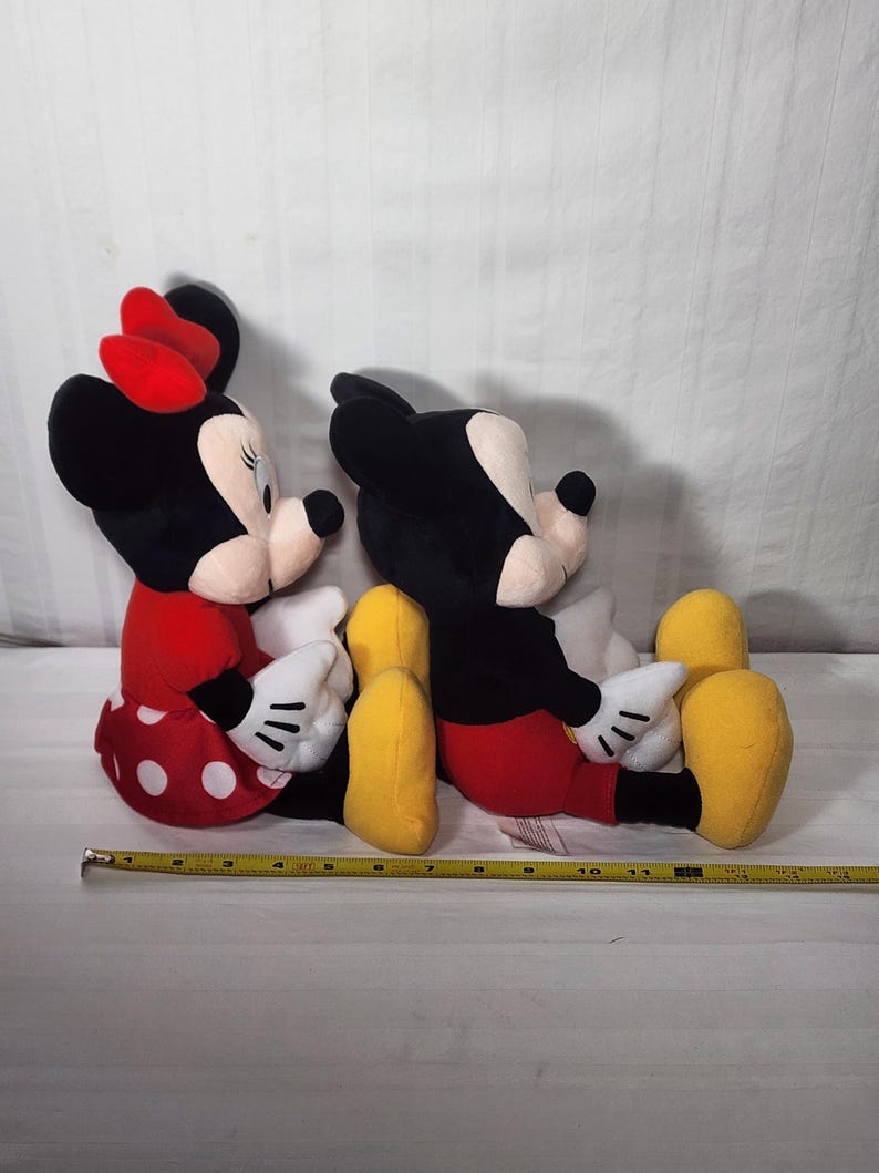 Pu&ograve; includere: Due bambole di peluche dei personaggi Disney, Minnie Mouse e Topolino, sono sedute fianco a fianco. Minnie indossa un vestito rosso a pois bianchi e un fiocco rosso. Topolino indossa pantaloncini rossi. Entrambi hanno orecchie nere e guanti bianchi.