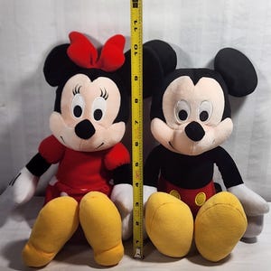 Pu&ograve; includere: Due personaggi Disney di peluche, Topolino e Minnie, sono esposti fianco a fianco. Minnie indossa un vestito e un fiocco rossi, mentre Topolino ha pantaloncini rossi. Un metro a nastro mostra che i giocattoli sono alti circa 28 cm.