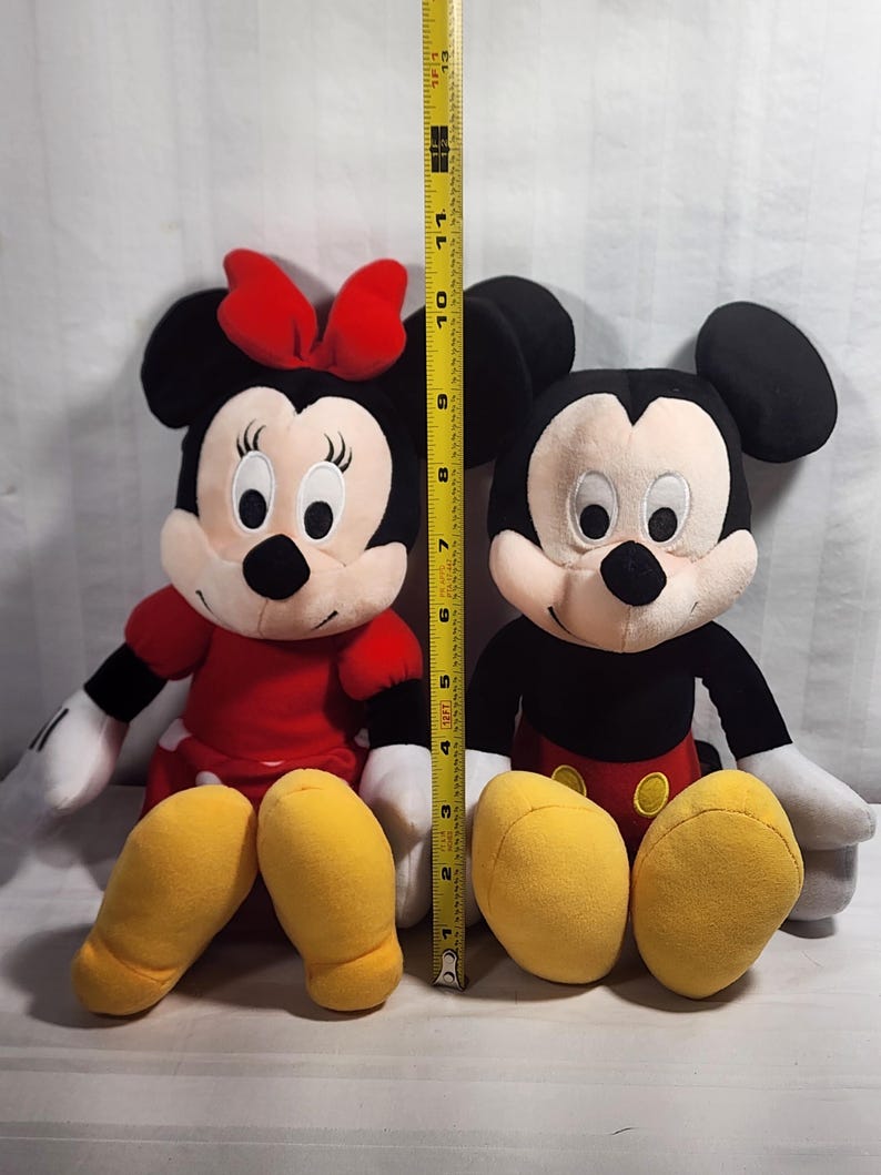 Pu&ograve; includere: Due personaggi Disney di peluche, Topolino e Minnie, sono esposti fianco a fianco. Minnie indossa un vestito e un fiocco rossi, mentre Topolino ha pantaloncini rossi. Un metro a nastro mostra che i giocattoli sono alti circa 28 cm.