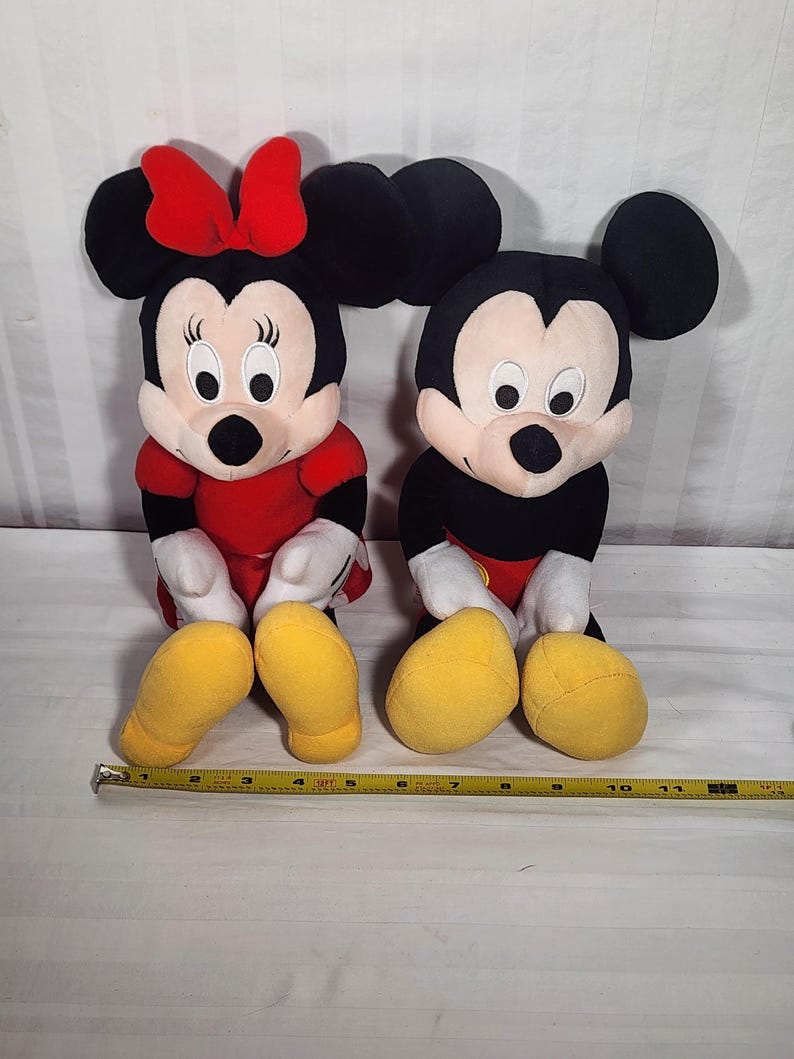 Pu&ograve; includere: Due bambole di peluche dei personaggi Disney, Minnie Mouse e Topolino, sono sedute fianco a fianco. Minnie indossa un vestito e un fiocco rossi, mentre Topolino ha pantaloncini rossi. Entrambi hanno orecchie nere, guanti bianchi e scarpe gialle.