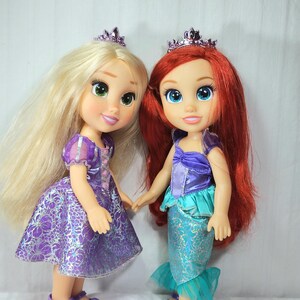 May include: Two dolls of Disney princesses, one with long blonde hair and a purple dress, the other with red hair and a mermaid tail. Both dolls wear crowns and hold hands. The dolls are standing in front of a white background.