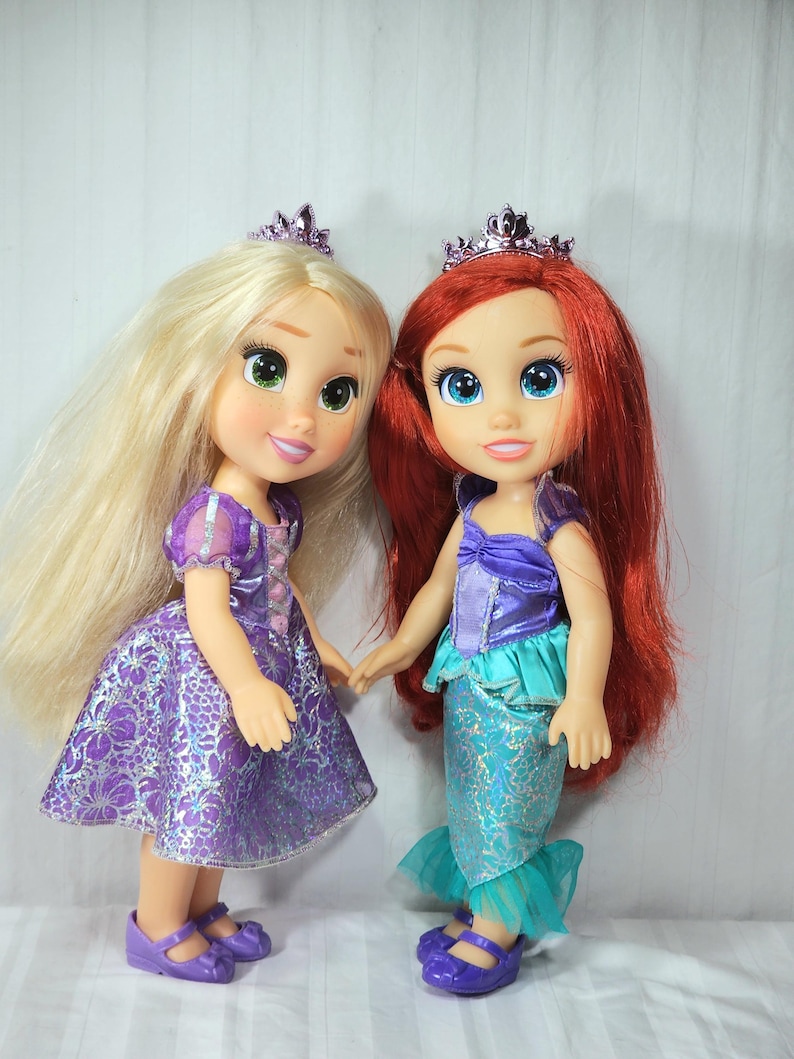 May include: Two dolls of Disney princesses, one with long blonde hair and a purple dress, the other with red hair and a mermaid tail. Both dolls wear crowns and hold hands. The dolls are standing in front of a white background.