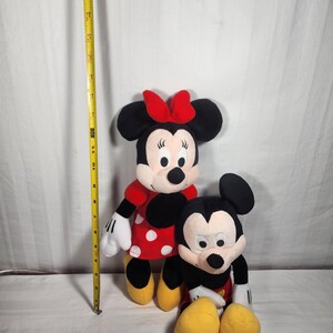 Pu&ograve; includere: Sono raffigurate due bambole di peluche dei personaggi Disney, Minnie Mouse e Topolino. Minnie Mouse indossa un vestito rosso a pois bianchi e un fiocco rosso. Topolino indossa pantaloncini neri, guanti bianchi e scarpe gialle. Un metro a nastro giallo &egrave; sullo sfondo.
