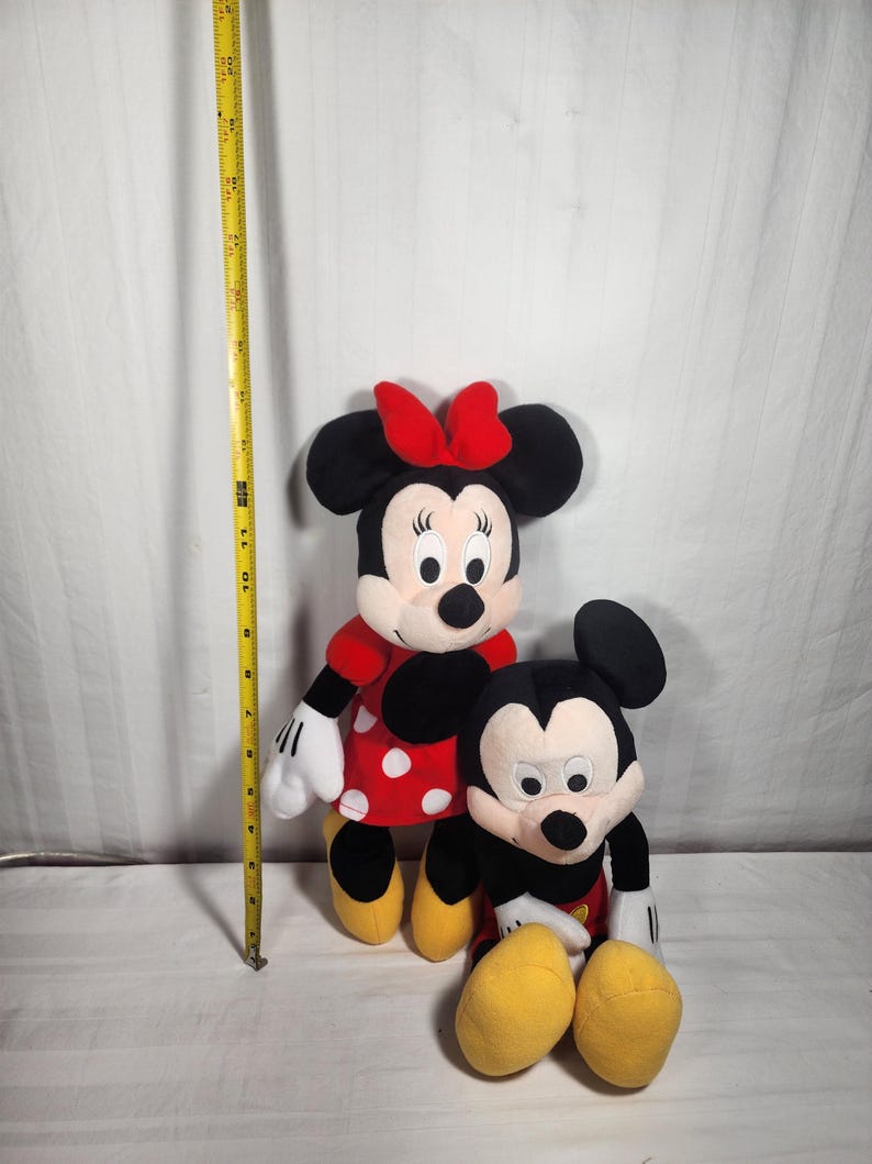Pu&ograve; includere: Sono raffigurate due bambole di peluche dei personaggi Disney, Minnie Mouse e Topolino. Minnie Mouse indossa un vestito rosso a pois bianchi e un fiocco rosso. Topolino indossa pantaloncini neri, guanti bianchi e scarpe gialle. Un metro a nastro giallo &egrave; sullo sfondo.