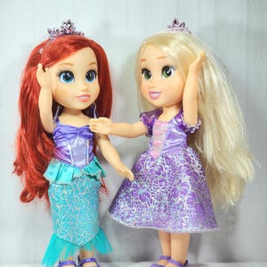 May include: Two dolls, one with red hair and a mermaid-style skirt, the other with blonde hair and a purple dress. Both dolls wear tiaras and have large, expressive eyes. The dolls are standing side-by-side, with one doll's arm touching the other.