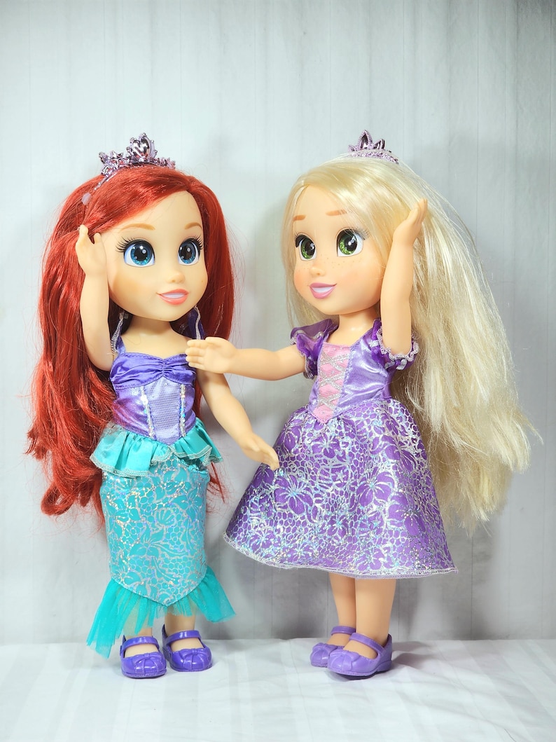 May include: Two dolls, one with red hair and a mermaid-style skirt, the other with blonde hair and a purple dress. Both dolls wear tiaras and have large, expressive eyes. The dolls are standing side-by-side, with one doll's arm touching the other.