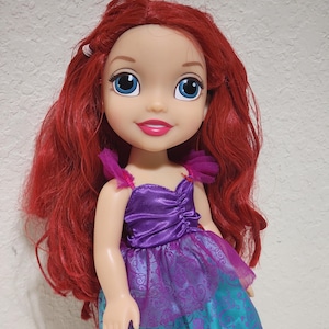 Puede incluir: Mu&ntilde;eca con cabello rojo y ojos azules, vestida con un vestido morado y turquesa. La mu&ntilde;eca tiene una expresi&oacute;n sonriente y es un personaje de una pel&iacute;cula de animaci&oacute;n infantil.