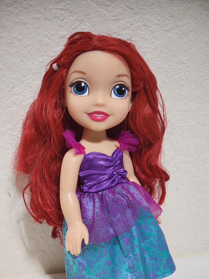 Puede incluir: Mu&ntilde;eca con cabello rojo y ojos azules, vestida con un vestido morado y turquesa. La mu&ntilde;eca tiene una expresi&oacute;n sonriente y es un personaje de una pel&iacute;cula de animaci&oacute;n infantil.