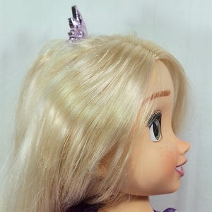 May include: A doll with long, blonde hair and a purple tiara. The doll has a fair complexion, freckles, and is wearing a purple dress. The doll's eyes are open, and it is looking to the side.