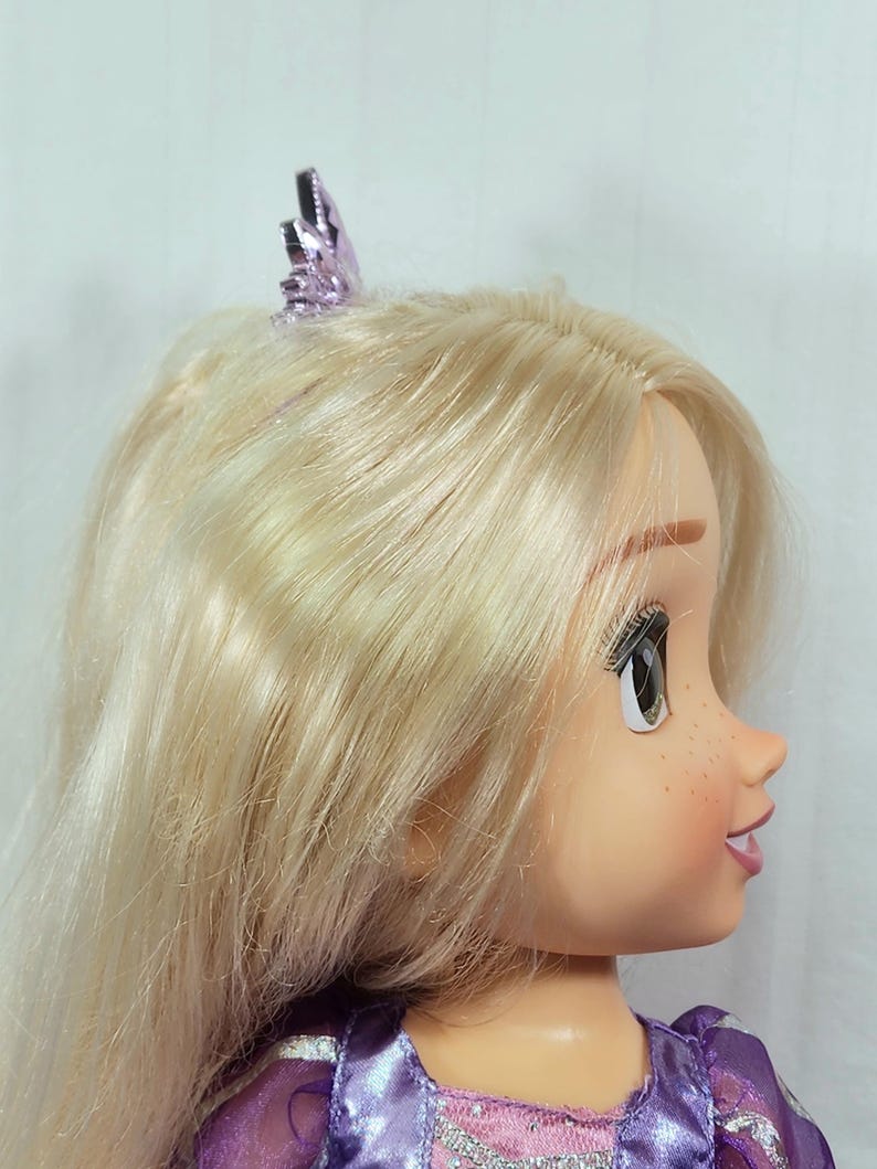 May include: A doll with long, blonde hair and a purple tiara. The doll has a fair complexion, freckles, and is wearing a purple dress. The doll's eyes are open, and it is looking to the side.