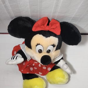 May include: A plush Minnie Mouse doll. The doll has large black ears, a red bow, and a red dress with white polka dots. The doll's face is cream-colored with large eyes and a black nose. The doll's feet are yellow, and it has white gloves.