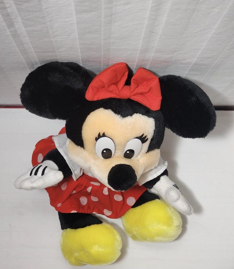 May include: A plush Minnie Mouse doll. The doll has large black ears, a red bow, and a red dress with white polka dots. The doll's face is cream-colored with large eyes and a black nose. The doll's feet are yellow, and it has white gloves.
