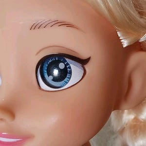 Op de afbeelding: Close-up van het gezicht van een pop met blauwe ogen en blond haar. De pop heeft een geschilderde glimlach en wimpers.