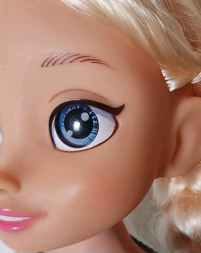 Op de afbeelding: Close-up van het gezicht van een pop met blauwe ogen en blond haar. De pop heeft een geschilderde glimlach en wimpers.