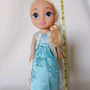 Op de afbeelding: Een blonde pop die een lichtblauwe jurk draagt met een sneeuwvlokkenpatroon. De pop heeft blauwe ogen en een roze mond. De pop is ongeveer 30 cm lang.